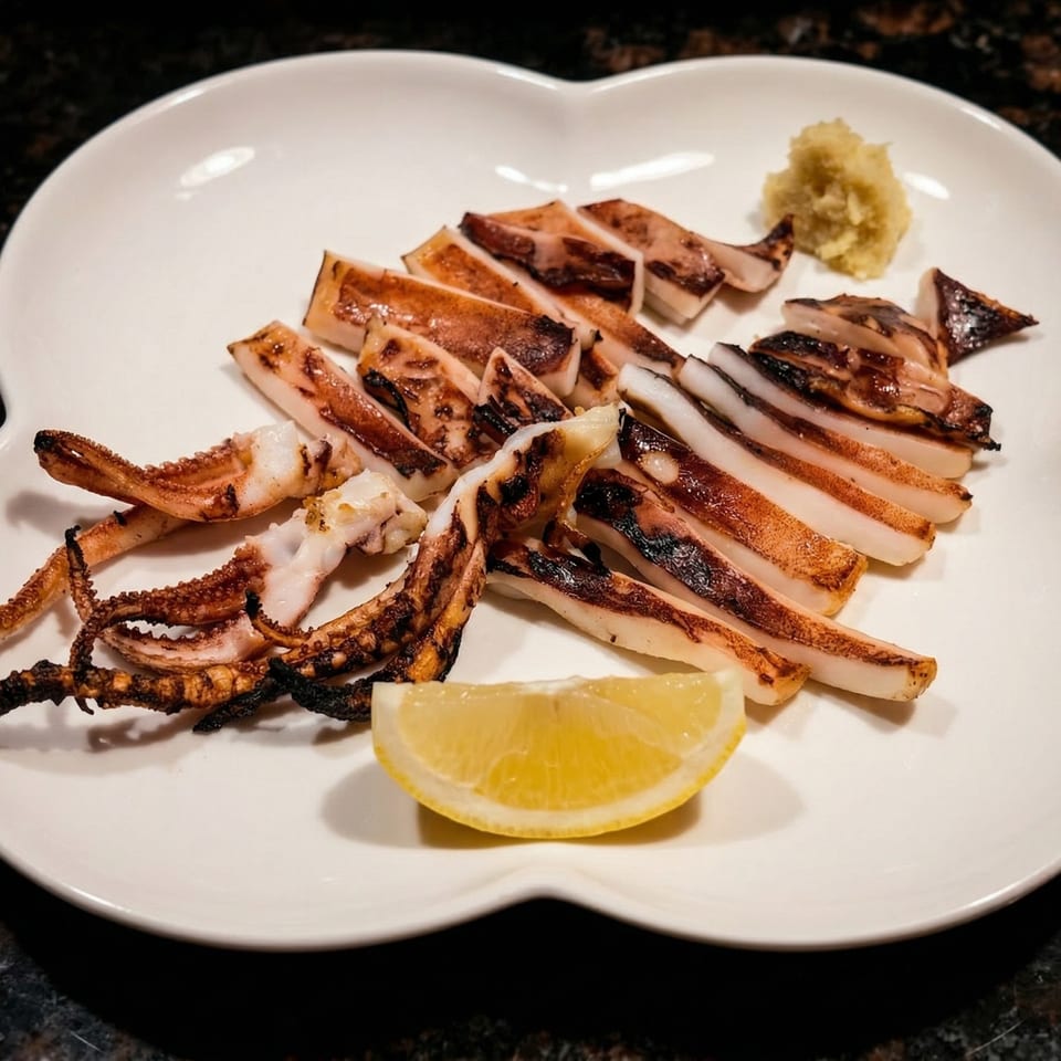 Grilled squid with tentacles and body slices, served with a lemon wedge and ginger on a white plate.