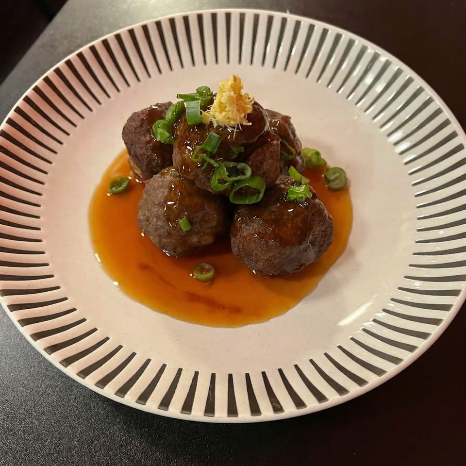 Meatballs in a rich brown sauce, garnished with green onions and shredded ginger on a striped plate.