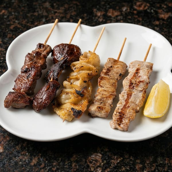 Grilled skewers of beef, mushrooms, chicken skin, and pork, with a lemon wedge on a white plate.