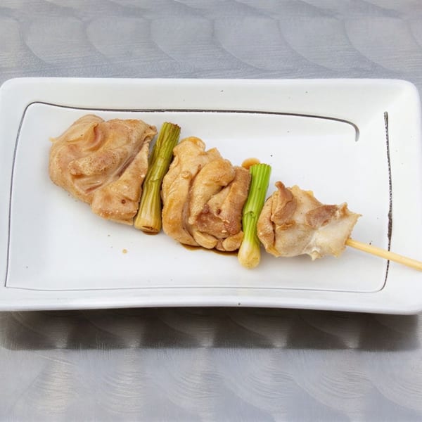 A yakitori skewer with chicken thigh and green onions on a white rectangular plate.